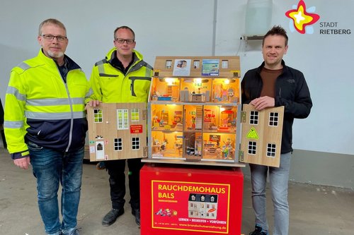 Die genannten Personen schauen in die Kamera. Sie stehen und präsentieren das Rauchdemohaus, das so groß ist wie ein Puppenhaus.