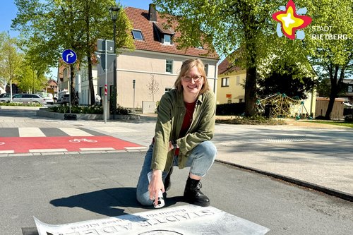 Eine Frau kniet im Sonnenschein auf der Straße und sprüht mit weißer Farbe einen Schriftzug auf den Asphalt.