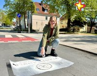 Eine Frau kniet im Sonnenschein auf der Straße und sprüht mit weißer Farbe einen Schriftzug auf den Asphalt.
