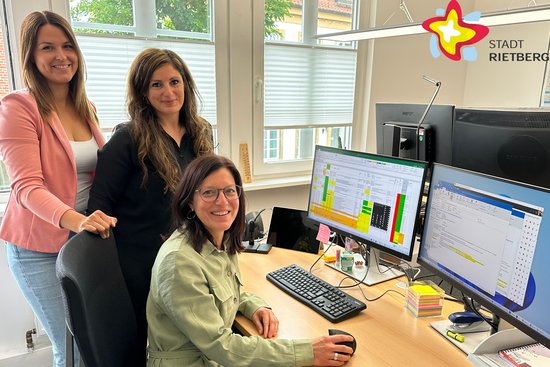 Drei Frauen stehen hinter einem Schreibtisch mit zwei Computerbildschirmen und lächeln in die Kamera.