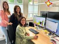 Drei Frauen stehen hinter einem Schreibtisch mit zwei Computerbildschirmen und lächeln in die Kamera.