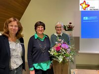 Die drei genannten Frauen stehen in der Friedhofskapelle und schauen frontal in die Kamera. Frau Peters hält einen Blumenstrauß in der Hand.