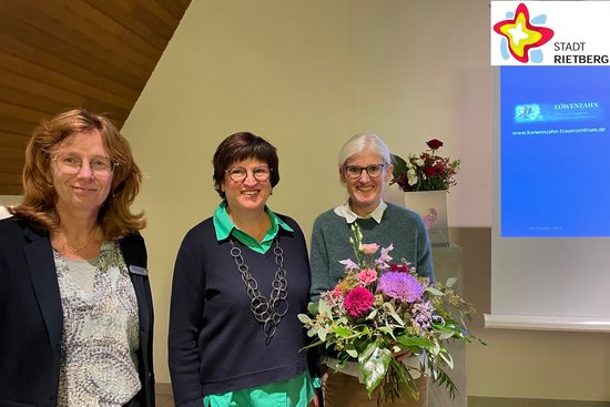 Die drei genannten Frauen stehen in der Friedhofskapelle und schauen frontal in die Kamera. Frau Peters hält einen Blumenstrauß in der Hand.