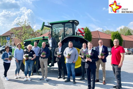 Mehrere Akteure haben sich vor einem Traktor auf dem Parkplatz neben dem Schnäppchen-Markt in Rietberg positioniert, um mit Blumen in der Hand auf die anstehende Wirtschaftsschau hinzuweisen.