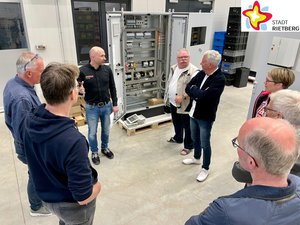 Elektrotechnikermeister Henryk Vogel steht mit  mehreren interessierten Zuhörern in einer Werkstatt vor einem geöffneten Schaltschrank.