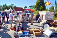 Bei strahlendem Sonnenschein betrachten mehrere Menschen den auf einem Parkplatz aufgebauten Trödel eines Mannes, der einen geöffneten Schirm in Regenbogenfarben über seinem Kopf hält.