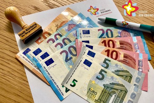 Mehrere Euro-Scheine liegen aufgefächert auf einem weißen Blatt Papier, daneben ein Stempel und ein Kugelschreiber mit der Aufschrift "Stadt Rietberg"