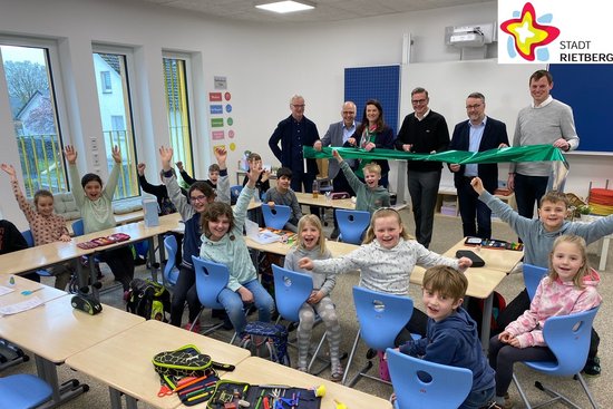 Fünf Männer und eine Frau stehen im Klassenraum der neuen Emsschule vor der Tafel und schneiden ein Band durch. Im Vordergrund sitzen Drittklässler an ihren Tischen. Alle schauen in die Kamera.