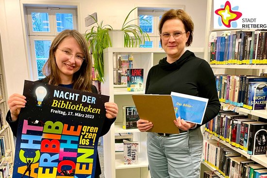 Jennifer Bader vom Bibliotheksteam und Moderatorin Annette Röwekamp stehen zwischen Bücherregalen in der Stadtbibliothek und zeigen Werbeplakat und Rätselkarten für das Biblio-Thekenquiz am 17. März.