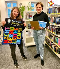 Jennifer Bader vom Bibliotheksteam und Moderatorin Annette Röwekamp stehen zwischen Bücherregalen in der Stadtbibliothek und zeigen Werbeplakat und Rätselkarten für das Biblio-Thekenquiz am 17. März.