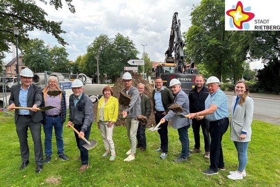11 Männer und Frauen stehen auf einer Wiese und schauen in die Kamera. Einige haben einen Bauhelm auf dem Kopf und halten Spaten mit Sand in der Hand. Im Hintergrund arbeitet ein Bagger.