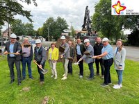 11 Männer und Frauen stehen auf einer Wiese und schauen in die Kamera. Einige haben einen Bauhelm auf dem Kopf und halten Spaten mit Sand in der Hand. Im Hintergrund arbeitet ein Bagger.