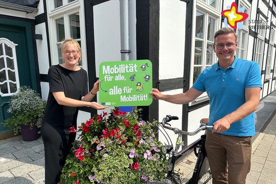 Rietbergs Mobilitätsbeauftragte Wibke Kordtomeikel und Bürgermeister Andreas Sunder mit einem Fahrrad stehen mit einem Werbeschild für die Europäische Mobilitätswoche vor einem Blumenkübel.