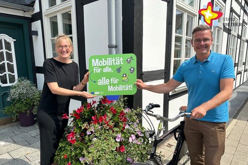 Rietbergs Mobilitätsbeauftragte Wibke Kordtomeikel und Bürgermeister Andreas Sunder mit einem Fahrrad stehen mit einem Werbeschild für die Europäische Mobilitätswoche vor einem Blumenkübel.