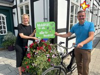 Rietbergs Mobilitätsbeauftragte Wibke Kordtomeikel und Bürgermeister Andreas Sunder mit einem Fahrrad stehen mit einem Werbeschild für die Europäische Mobilitätswoche vor einem Blumenkübel.