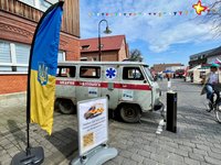 Ein beschossener, grauer, alter Rettungswagen steht auf einer Straße in Langenberg. Im Vordergrung ist ein Aufsteller mit ukrainischer Flagge zu sehen. Eine Radfahrerin fährt vorbei. In der Luft hängt eine Wimpelkette.