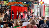 Kinder tanzen vor einem roten Vorhang auf einer kleinen Bühne und ein Publikum auf Stühlen sitzend schaut zu.