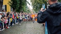 Kinder laufen über Kopfsteinpflaster, links und rechts gesäumt von weiteren Kindern und und Erwachsenen als Zuschauer.