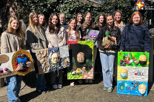 Die Jugendlichen und ihre Lehrerin stehen zusammen und zeigen ihre Kunstwerke aus recycelten Materialien. Zu sehen sind vier große Bilder, ein kleines Bild und eine Skulptur.