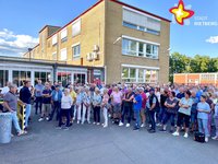 Die beiden genannten Personen und viele weitere Menschen stehen vor den Hallen der ehemaligen Firma Lear. Die Sonne scheint. Der Himmel ist blau.