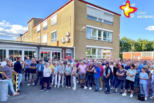 Die beiden genannten Personen und viele weitere Menschen stehen vor den Hallen der ehemaligen Firma Lear. Die Sonne scheint. Der Himmel ist blau.