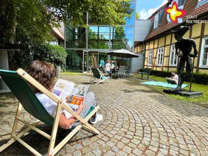 Das Bild zeigt eine Frau von hinten im Liegestuhl sitzend mit einem Buch in den Händen. Im Hintergrund sitzt eine Frau unter einem Sonnenschirm an einem Tisch.