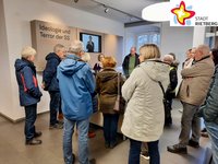 Interessiert stehen mehrere Menschen im Kreis um eine gläserne Vitrine herum und drehen der Kamera den Rücken zu.