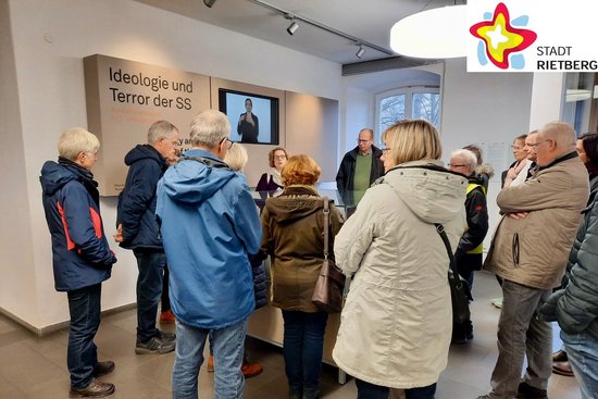 Interessiert stehen mehrere Menschen im Kreis um eine gläserne Vitrine herum und drehen der Kamera den Rücken zu.