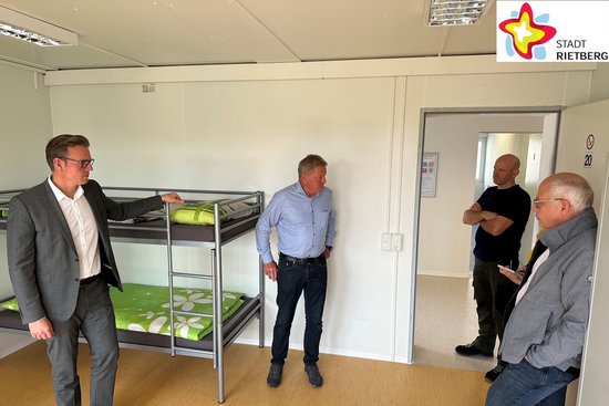 Vier Personen stehen in einem Raum einer Flüchtlingsunterkunft. Es ist ein Etagenbett zu sehen, eine Tür zum Flur steht auf.