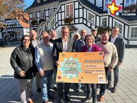 Einige Personen stehen vor dem Rathaus.
Sie schauen in die Kamera.
Sie halten einen großen Scheck hoch.