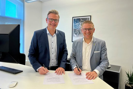 Die beiden Bürgermeister stehen an einem Schreibtisch und schauen in die Kamera. Sie haben Stifte in der Hand und sind dabei, einen Vertrag zu unterschreiben.