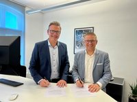 Die beiden Bürgermeister stehen an einem Schreibtisch und schauen in die Kamera. Sie haben Stifte in der Hand und sind dabei, einen Vertrag zu unterschreiben.