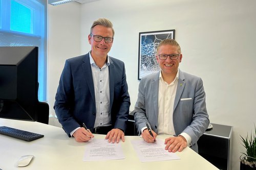 Die beiden Bürgermeister stehen an einem Schreibtisch und schauen in die Kamera. Sie haben Stifte in der Hand und sind dabei, einen Vertrag zu unterschreiben.