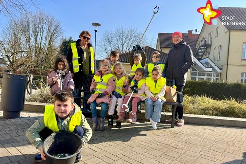 Kinder in gelben Warnwesten sitzen auf einer Bank, eingerahmt von zwei erwachsenen Frauen, und lächeln in die Kaerma. Im Vordergrund sitzt ein junge in der Hocke und hält einen Eimer in die Kamera.