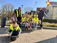 Kinder in gelben Warnwesten sitzen auf einer Bank, eingerahmt von zwei erwachsenen Frauen, und lächeln in die Kaerma. Im Vordergrund sitzt ein junge in der Hocke und hält einen Eimer in die Kamera.