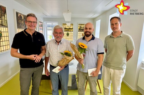Vier Männer stehen nebeneinander und lächeln in die Kamera. Die zwei Männer in der Mitte halten je einen Blumenstrauß.