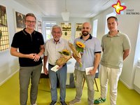 Vier Männer stehen nebeneinander und lächeln in die Kamera. Die zwei Männer in der Mitte halten je einen Blumenstrauß.