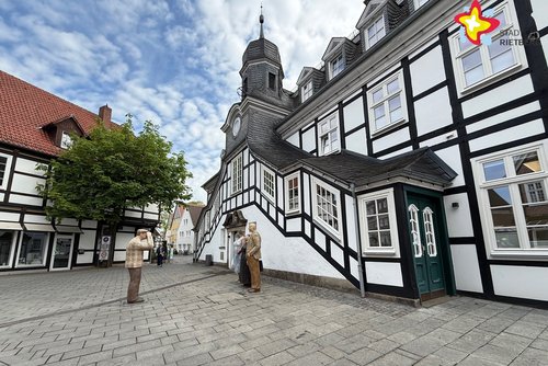 Das Bild zeigt das Rietberger Rathaus von der Seite. Davor stehen Alltagsmenschen-Skulpturen. Links blüht eine Kastanie.