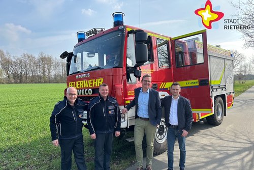 Die genannten Personen stehen vor einem Feuerwehrauto und schauen in die Kamera. Im Hintergrund ist eine grüne weitläufige Wiese zu erkennen. Der Himmel ist hellblau.