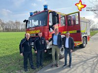 Die genannten Personen stehen vor einem Feuerwehrauto und schauen in die Kamera. Im Hintergrund ist eine grüne weitläufige Wiese zu erkennen. Der Himmel ist hellblau.