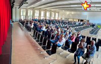 Vor der Bühne einer Aula sind die Stuhlreihen zur Hälfte besetzt, während im hinteren Teil weitere Frauen und Männer stehen. Links im Bild verdeckt der rote Vorhang die Bühne.