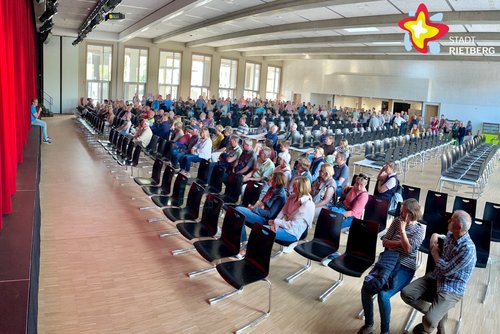 Vor der Bühne einer Aula sind die Stuhlreihen zur Hälfte besetzt, während im hinteren Teil weitere Frauen und Männer stehen. Links im Bild verdeckt der rote Vorhang die Bühne.