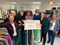 Die genannten Personen stehen inmitten von Kleidung im Geschäft und schauen in die Kamera. Sie halten einen großen Scheck in den Händen.