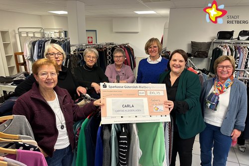 Die genannten Personen stehen inmitten von Kleidung im Geschäft und schauen in die Kamera. Sie halten einen großen Scheck in den Händen.