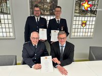 Die genannten Personen sitzen (zwei) und stehen in einem Raum im Rathaus und halten Urkunden in den Händen.