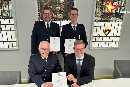 Die genannten Personen sitzen (zwei) und stehen in einem Raum im Rathaus und halten Urkunden in den Händen.