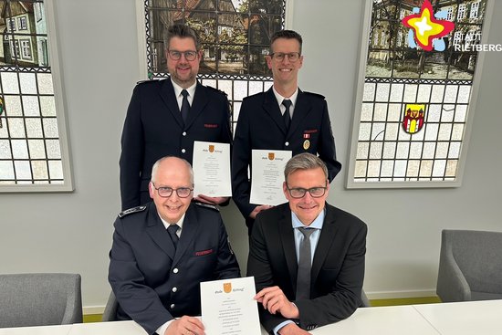 Die genannten Personen sitzen (zwei) und stehen in einem Raum im Rathaus und halten Urkunden in den Händen.