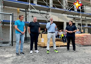 Vier Männer stehen nebeneinander vor dem Rohbau einer Baustelle. Der dritte von links hält ein Mikrofon in der Hand.