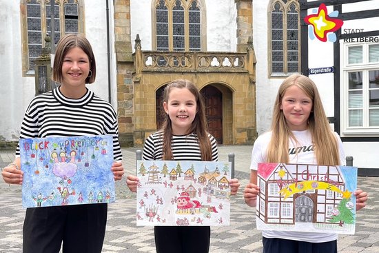 Inga Moorfeld, Lina Paulfeuerborn und Mara Südhaus stehen nebeneinander vor der Pfarrkirche und zeigen ihre selbst gemalten Bilder für die Adventszeit.