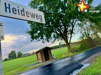 Ein Straßenschild mit der Aufschrift "Heideweg" ragt von links in das Bild, während im Hintergrund eine frisch asphaltierte Straße zu sehen ist.